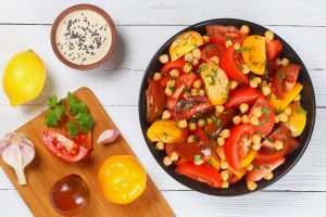 chickpea tomatoes salad with parsley in black bowl, homemade tahini sauce sprinkled with black sesame seeds and bottle of olive oil on white table, ingredients on chopping board, view from above